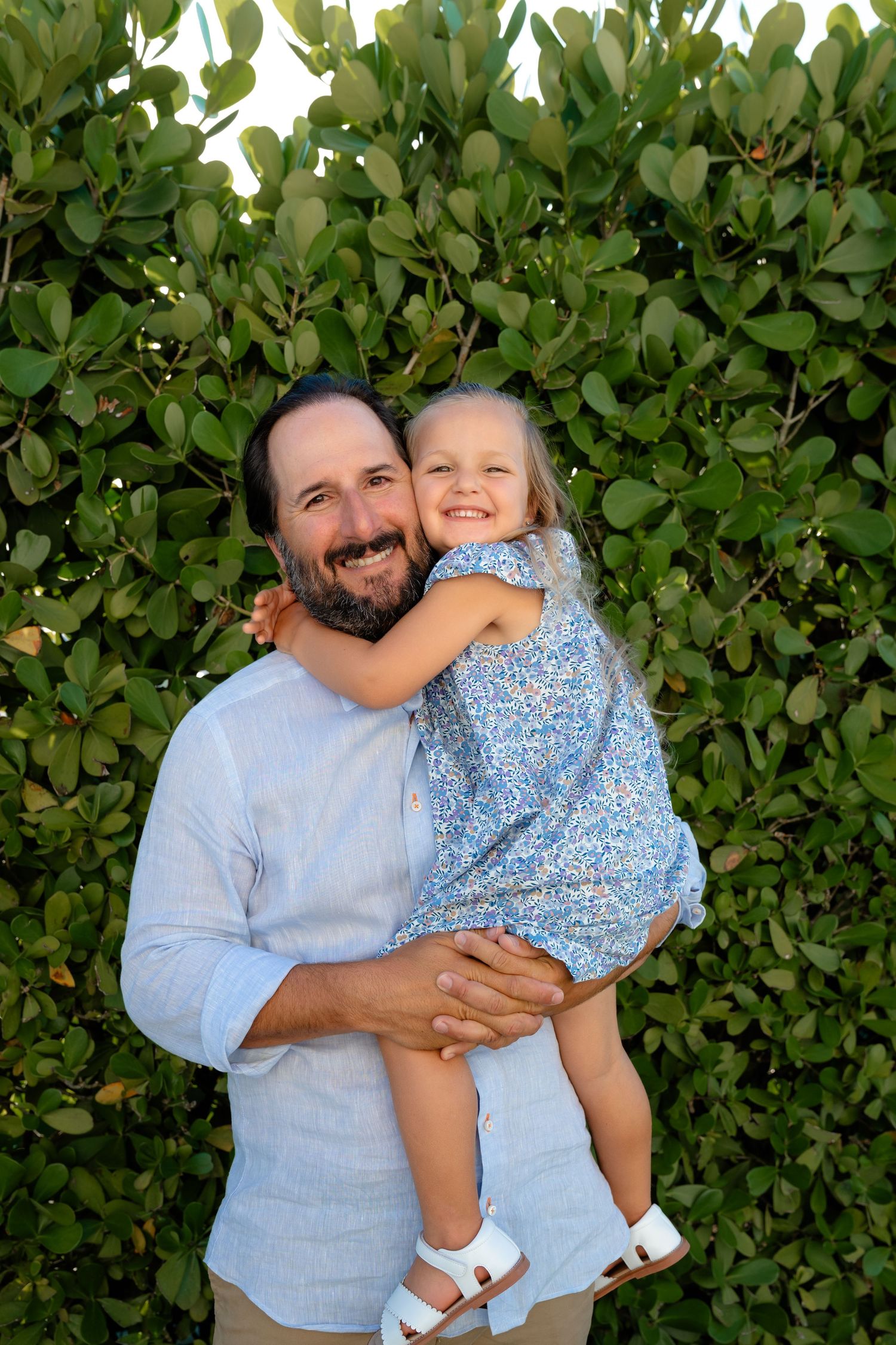 Smiling bearded man in blue shirt holding a happy young girl in floral dress outdoors near green foliage.