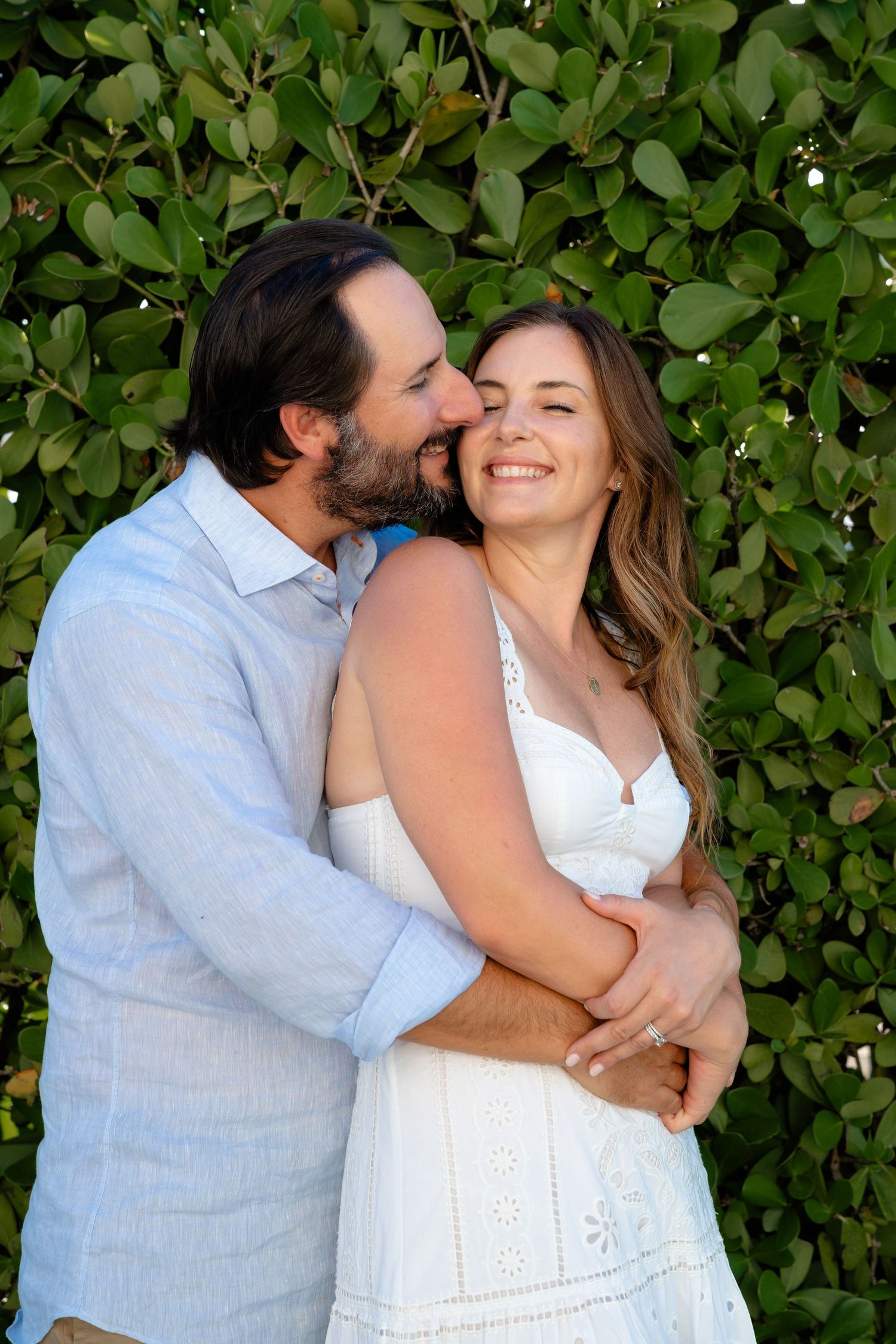 A couple embraces outdoors, man kissing woman's cheek as she smiles, both dressed in white and blue against green foliage.