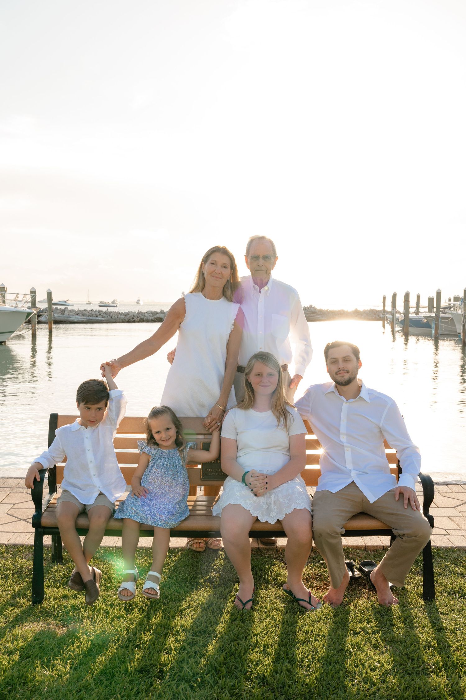 Family of six posing on a wooden bench by a marina at sunset, dressed in white and light blue outfits.