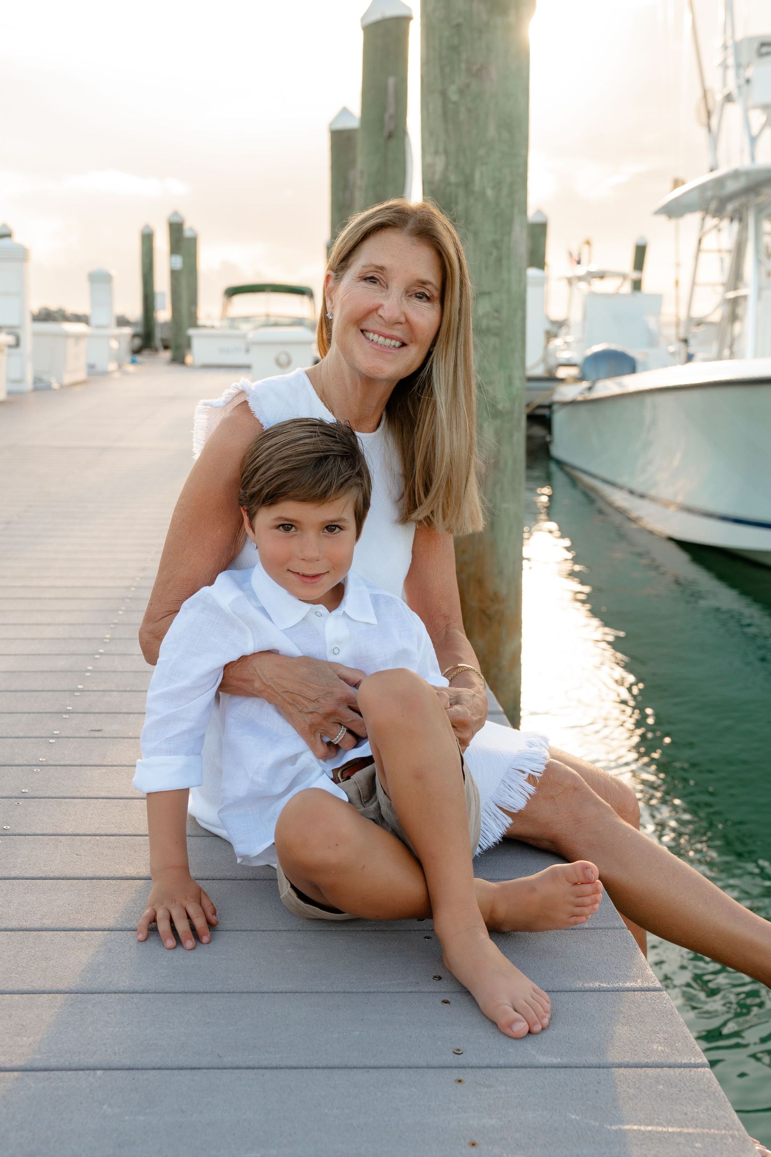 A smiling woman and young boy in white outfits sit together on a marina dock at sunset.