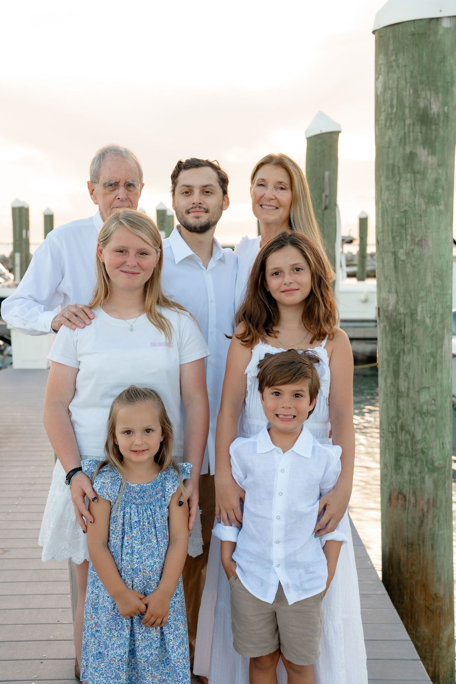 Multi-generational family of seven poses on a marina dock at sunset, dressed in white and blue coordinated outfits.