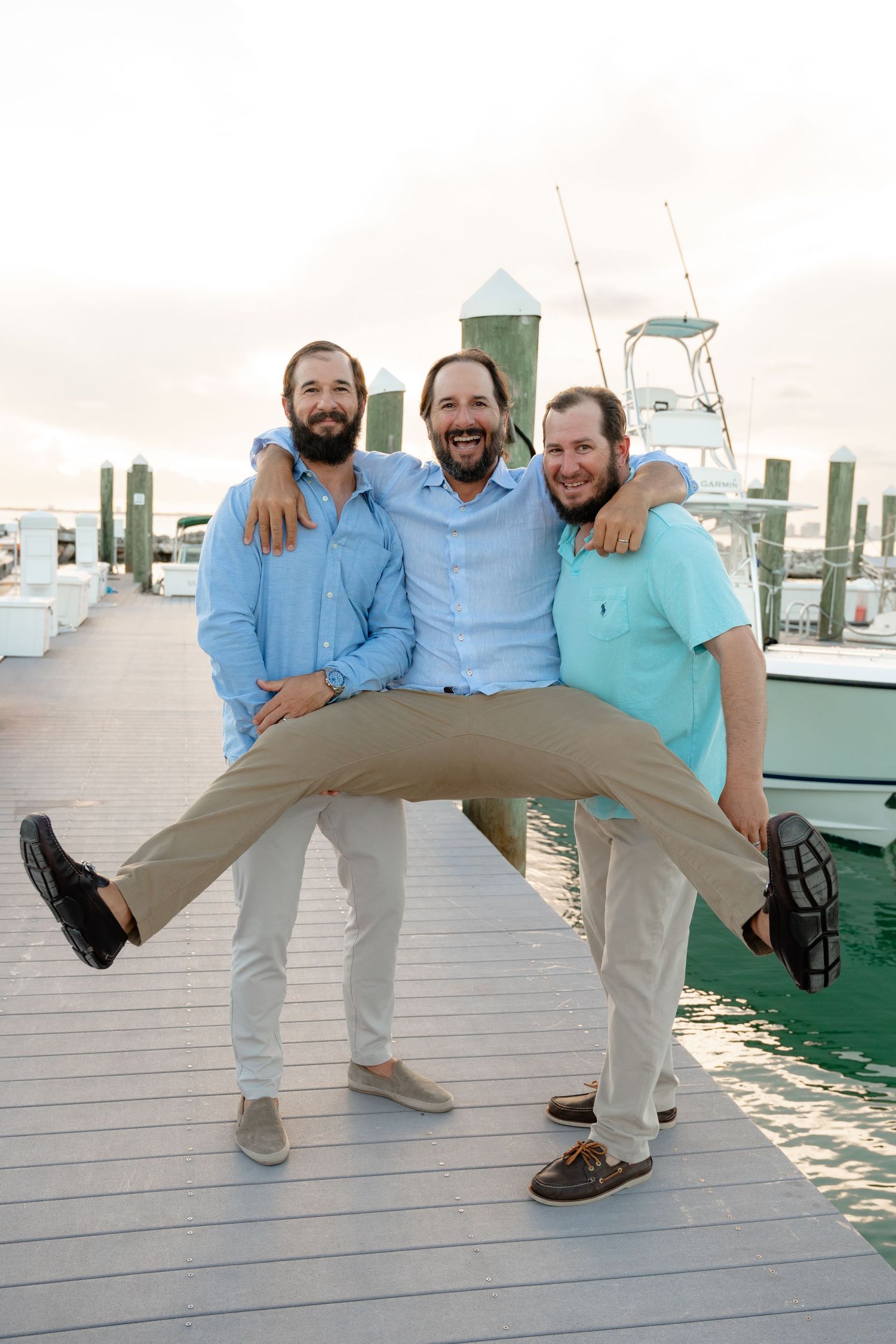 Three smiling men pose playfully on a marina dock, wearing casual pastel shirts with boats in the background.