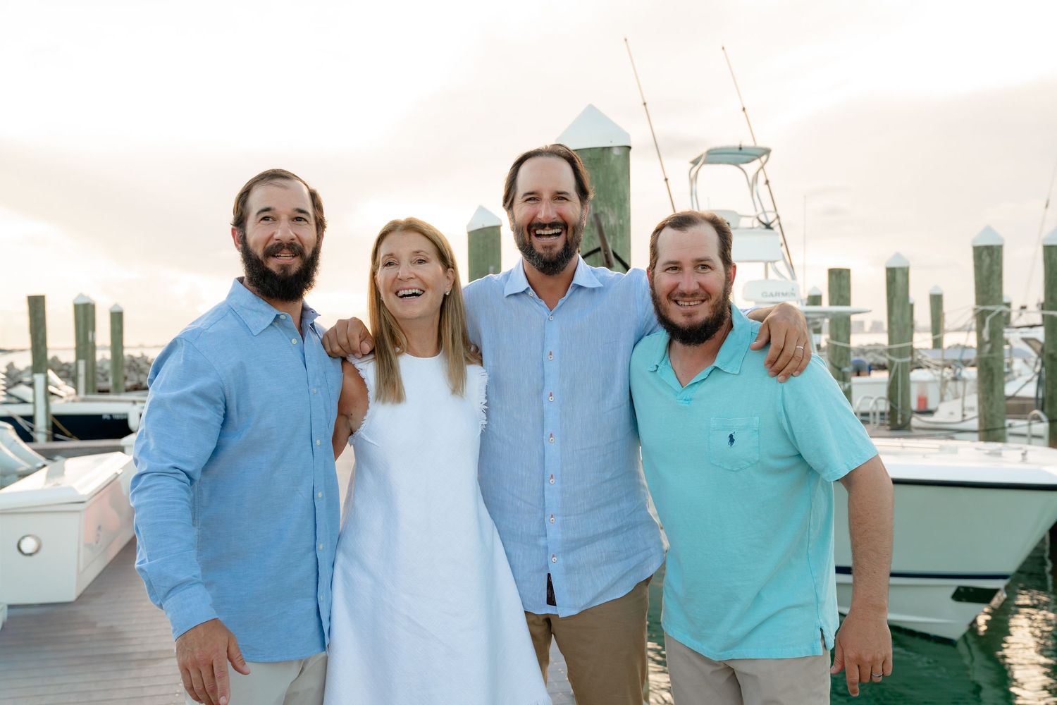 Four friends smiling together on a marina dock, dressed in casual summer attire near docked boats.