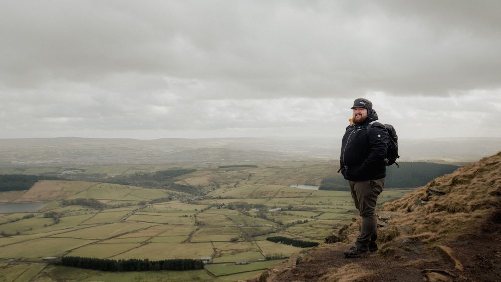 Hiker with backpack stands on rocky hilltop overlooking vast green patchwork valley under overcast sky.