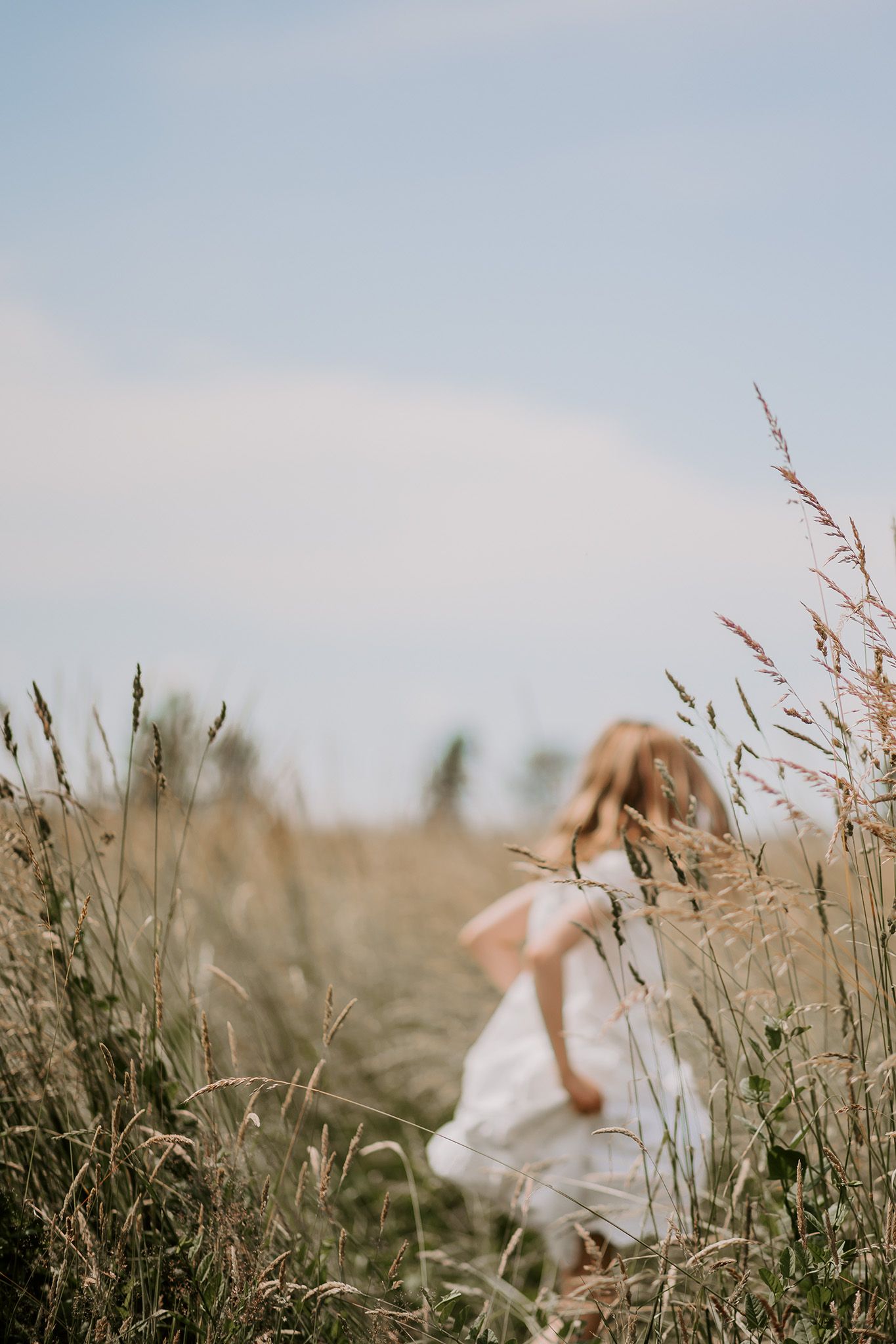 En person i en hvid kjole sidder blandt højt græs og vilde blomster i en drømmende udendørs setting. Familiefotograf Fyn