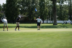 Golfers on a sunny green course near the flag, with trees and a house in the background.