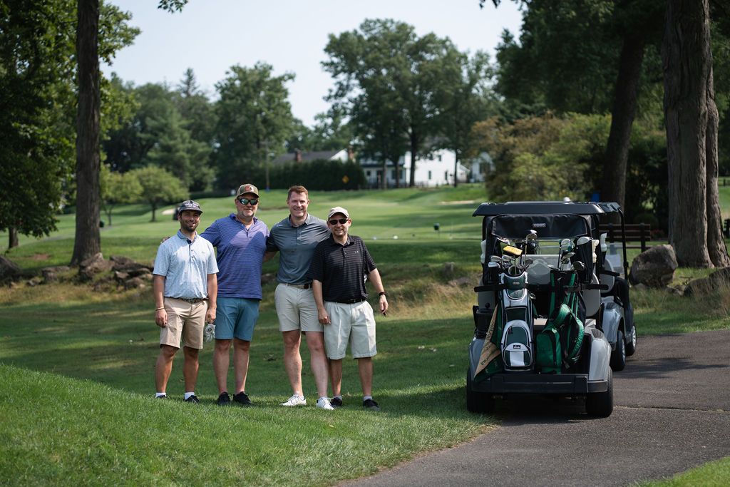 Four golfers pose together near a golf cart on a lush green golf course on a sunny day.
