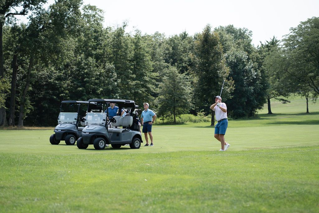 Golfers near golf carts on a lush green fairway surrounded by trees on a sunny day.