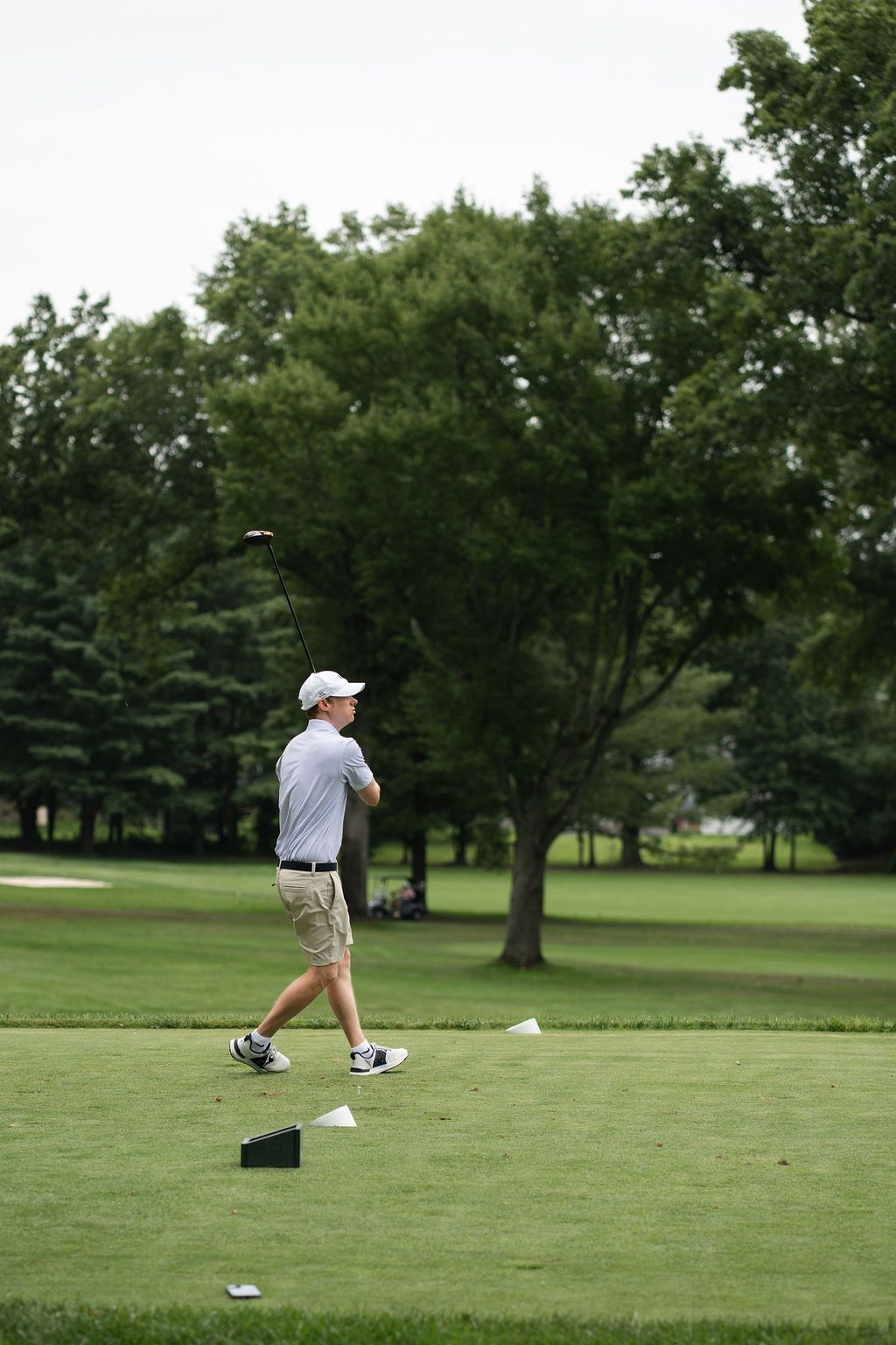 Young golfer in white cap and khaki shorts swings a club on a lush green golf course surrounded by trees.