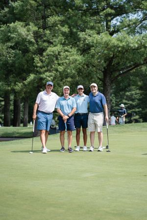 Four men posing together on a golf course green, holding putters, surrounded by tall pine trees on a sunny day.