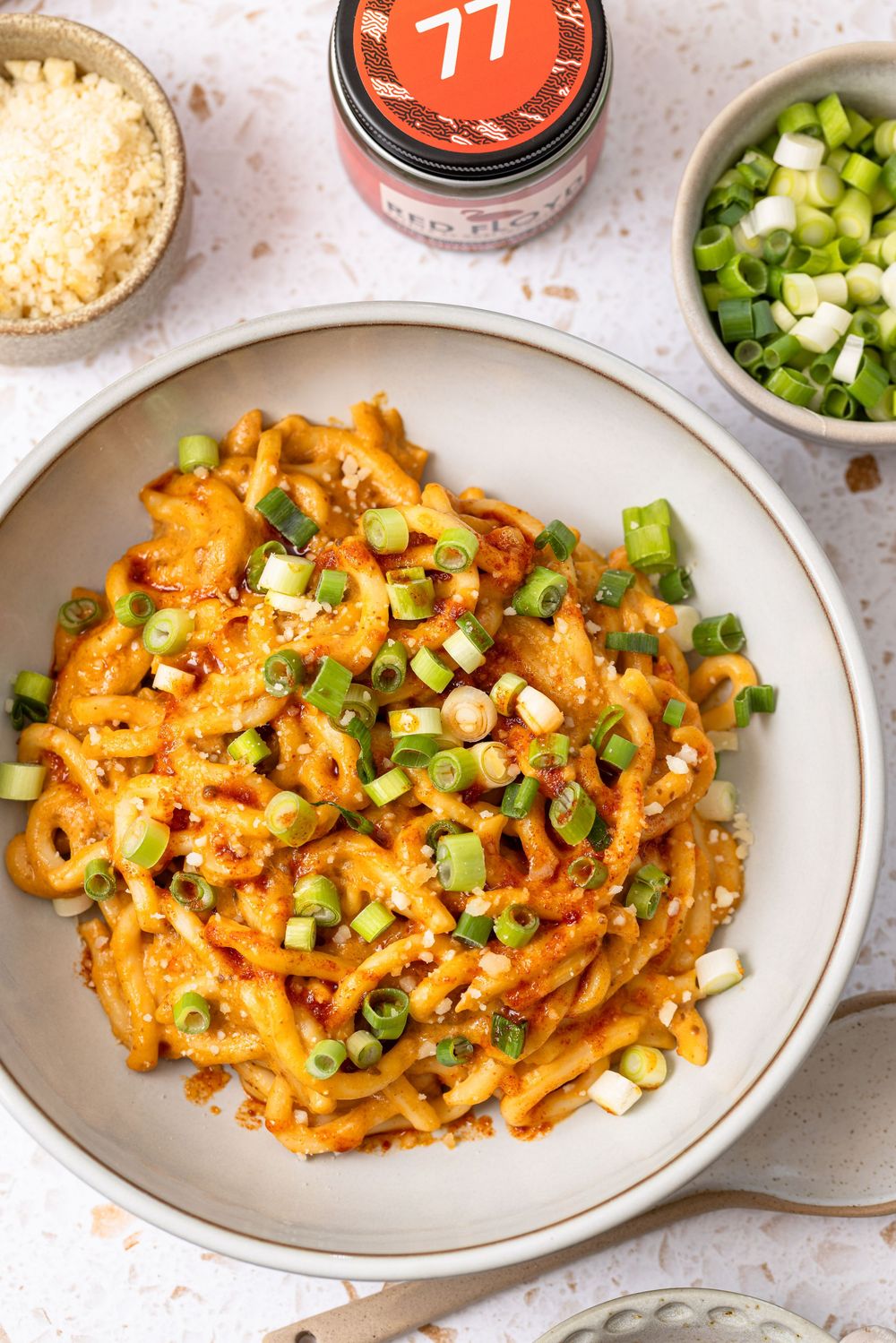 A plate of spicy red sauce pasta topped with fresh green onions, served with grated cheese and a jar of red flakes.