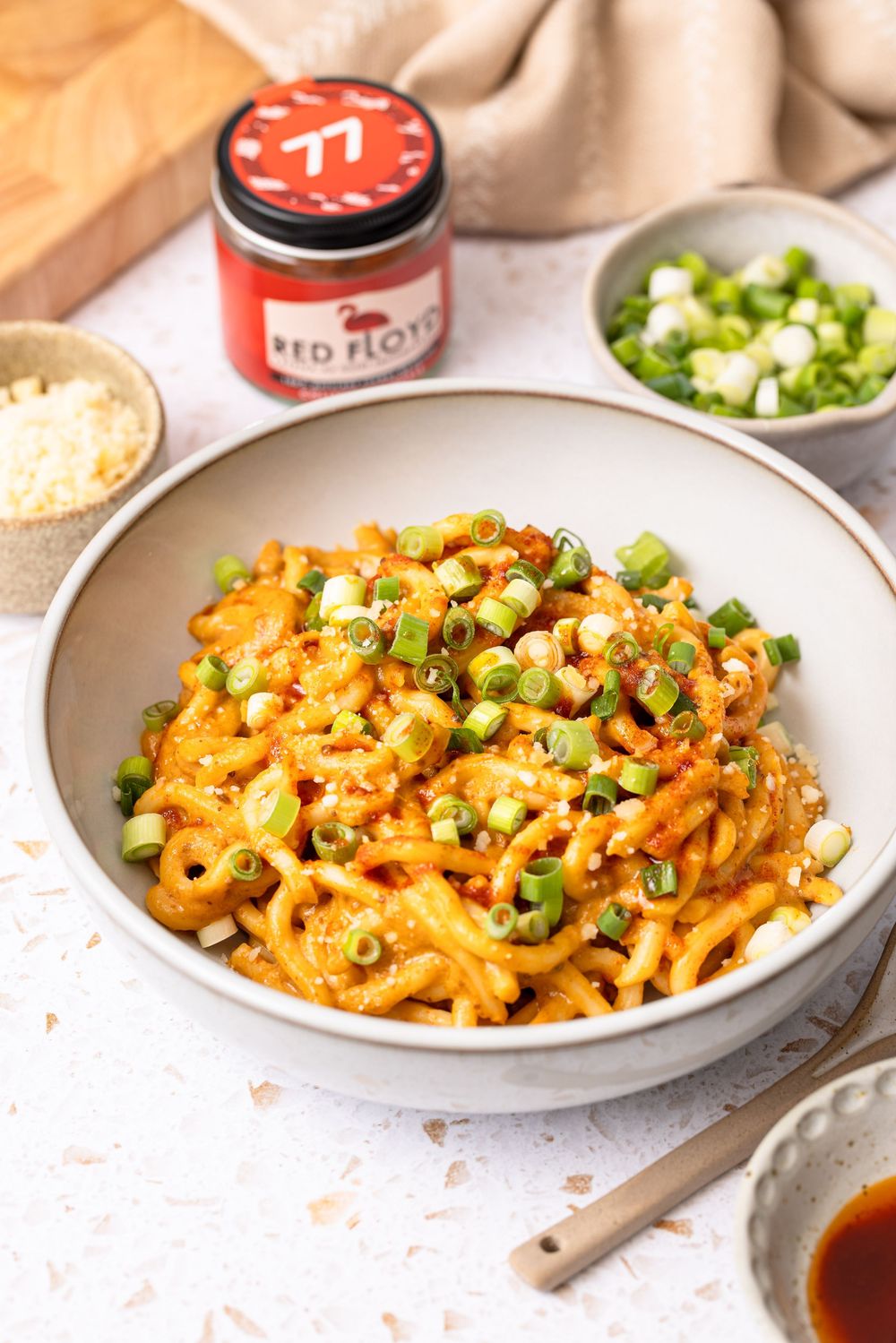 A bowl of spicy udon noodles topped with green onions, served with Red Floyd seasoning, cheese, and dipping sauce.
