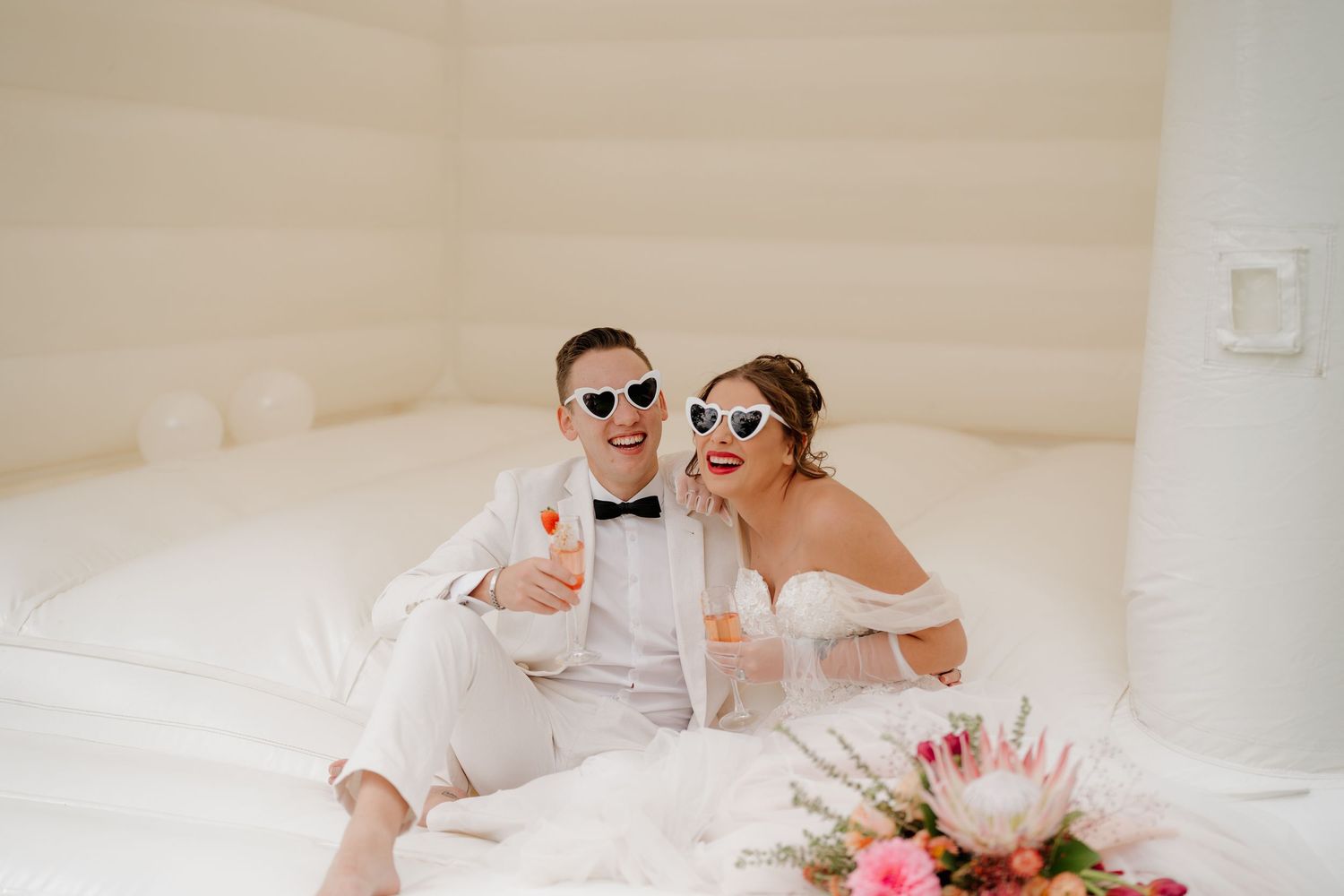 Bride and groom in sunglasses sitting in white bounce house, holding champagne flutes, with floral arrangement nearby.