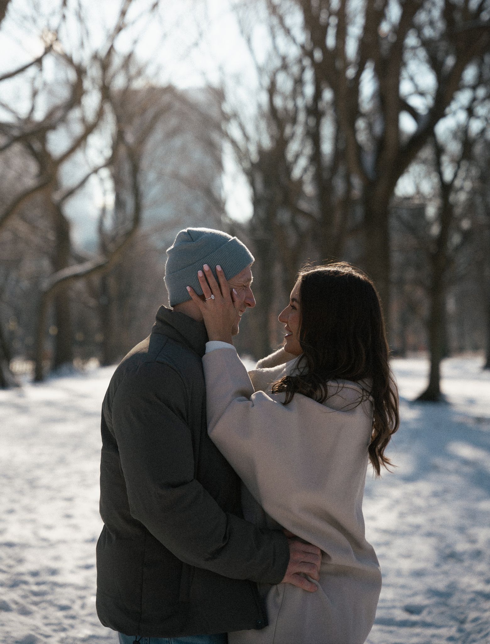 Couple embracing and smiling at each other in a snowy winter park, surrounded by bare trees on a sunny day.