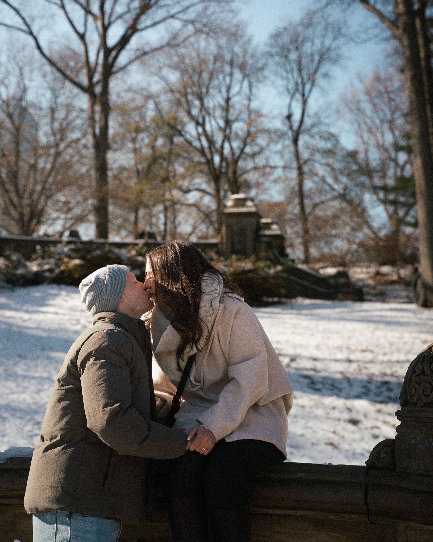 Couple sharing a romantic kiss in snowy Central Park, wearing winter coats, with bare trees and snow-covered ground behind them.