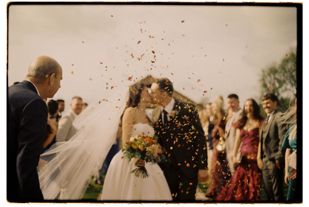 Newlywed couple sharing a kiss while guests throw colorful confetti during outdoor wedding ceremony.