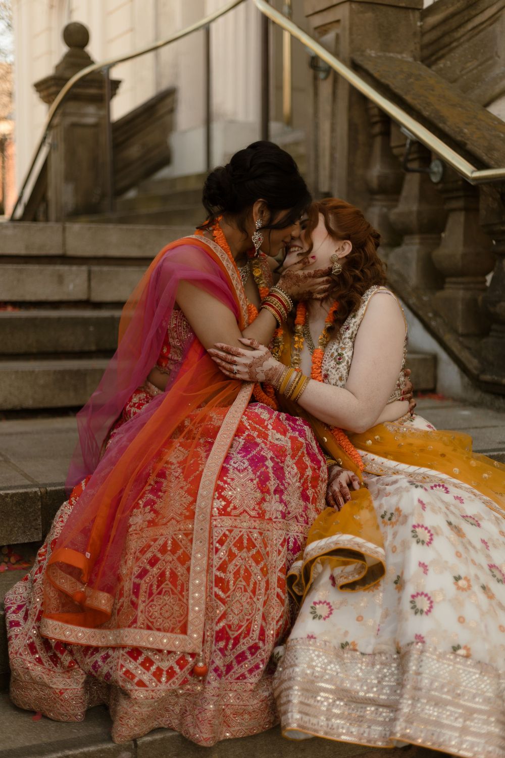 Two brides in vibrant Indian bridal lehenga outfits share a romantic kiss on stone steps.