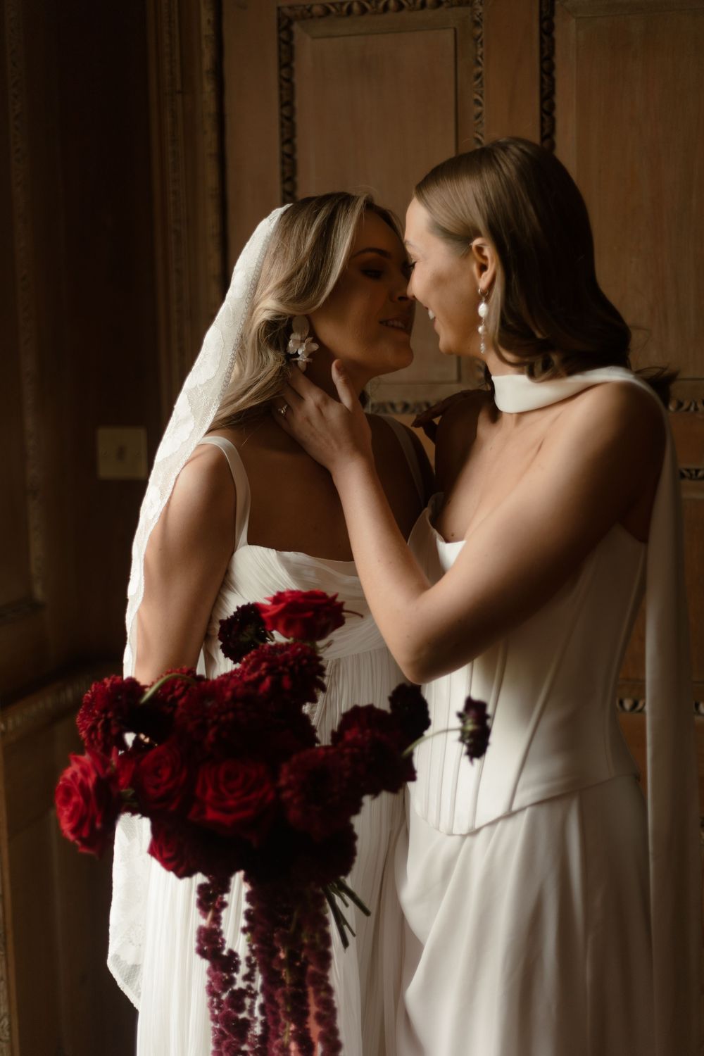 Two brides in white gowns share an intimate moment, one holding a cascading bouquet of deep red flowers.