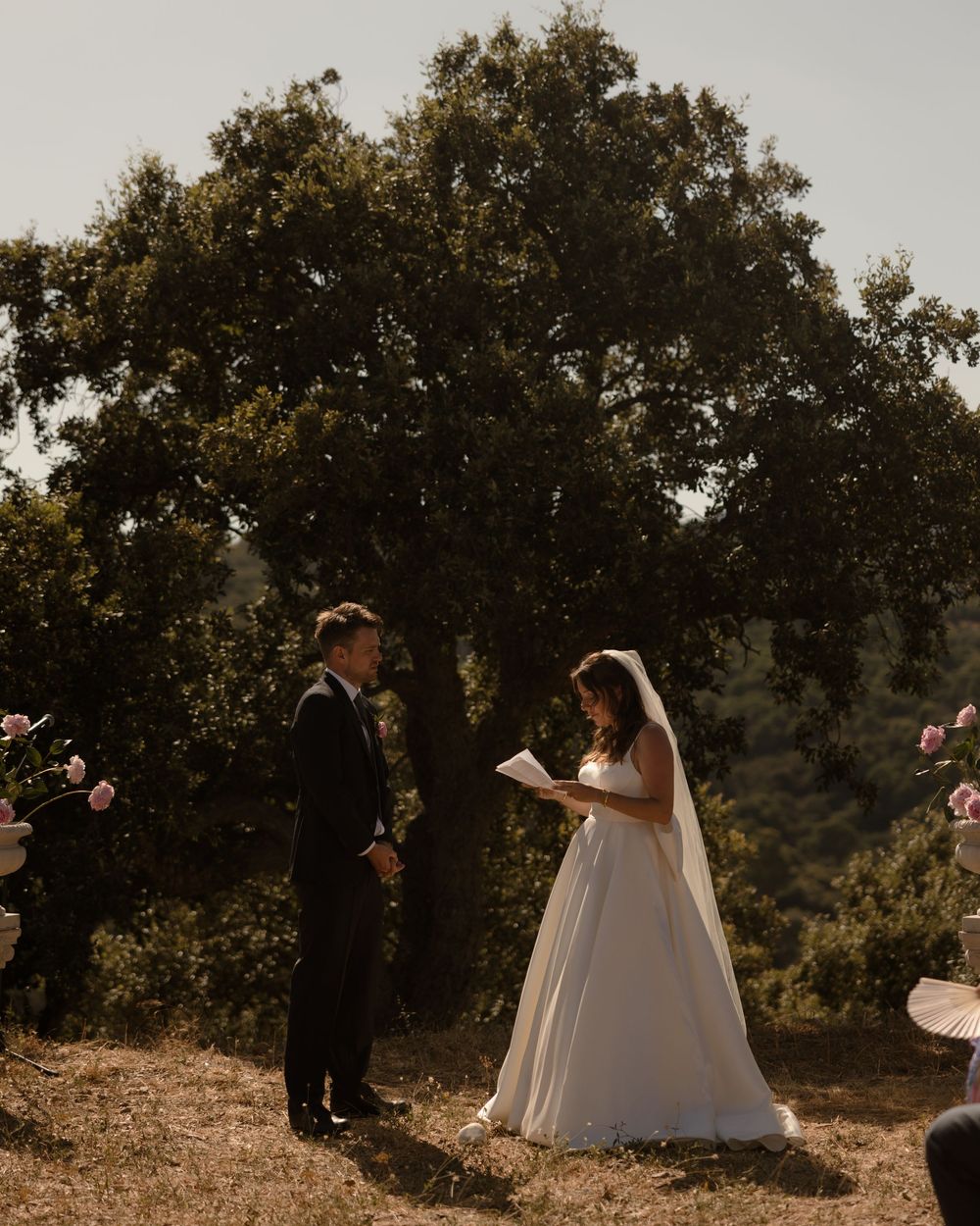 Bride reads vows to groom outdoors at rustic wedding ceremony surrounded by pink roses and lush green trees.