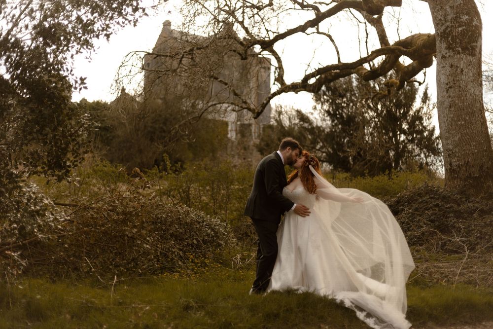 Bride and groom share romantic kiss in white wedding dress and suit before historic castle ruins outdoors.