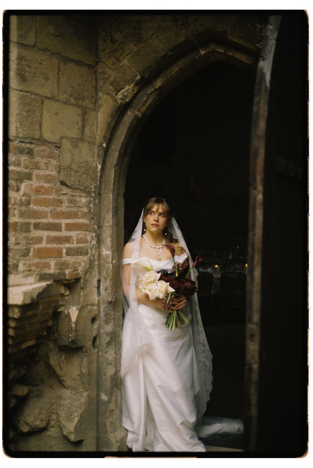 Bride in white gown and veil holding bouquet standing in stone archway doorway of historic castle venue.