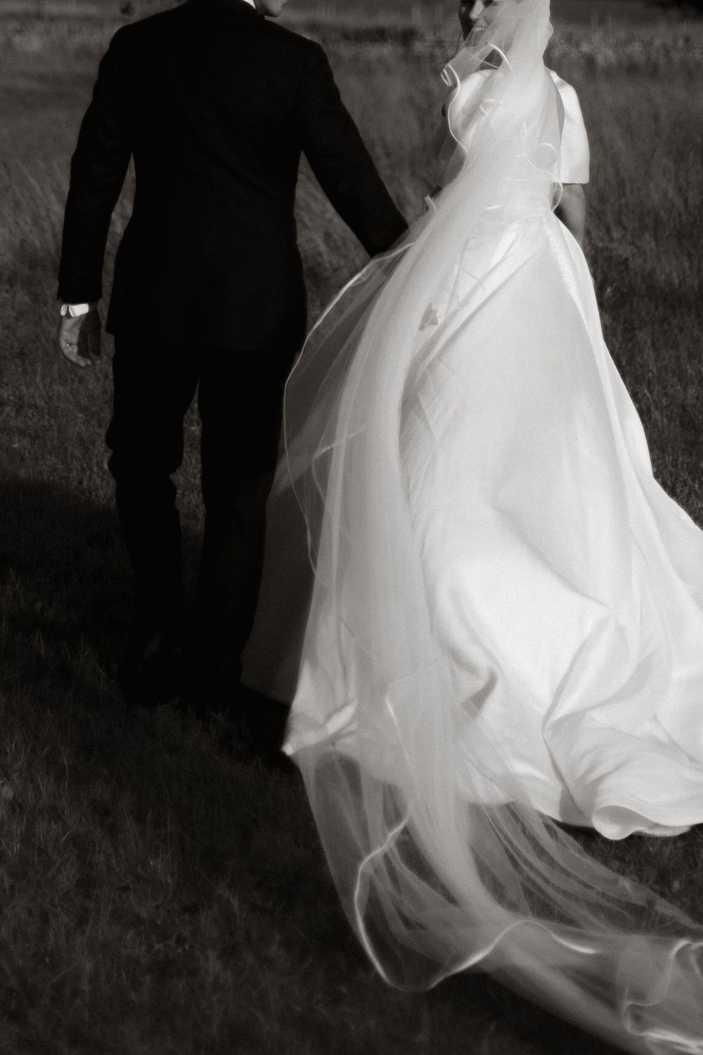 Black and white photo of a bride in flowing gown and groom in suit walking together across a grassy field.