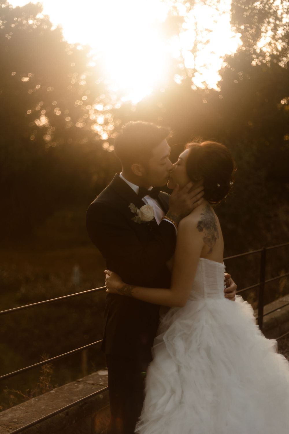 Bride and groom share a romantic kiss at golden hour sunset, with warm glowing light filtering through trees.