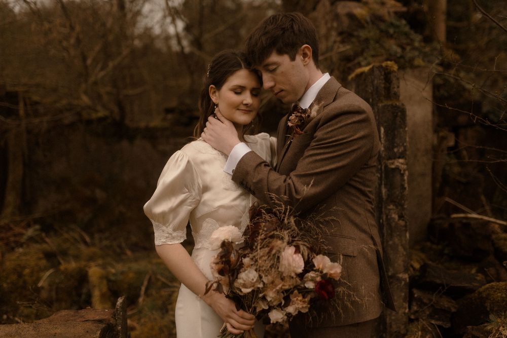 Bride and groom embrace tenderly in autumn woodland setting, she holds dried floral bouquet, he wears brown suit.