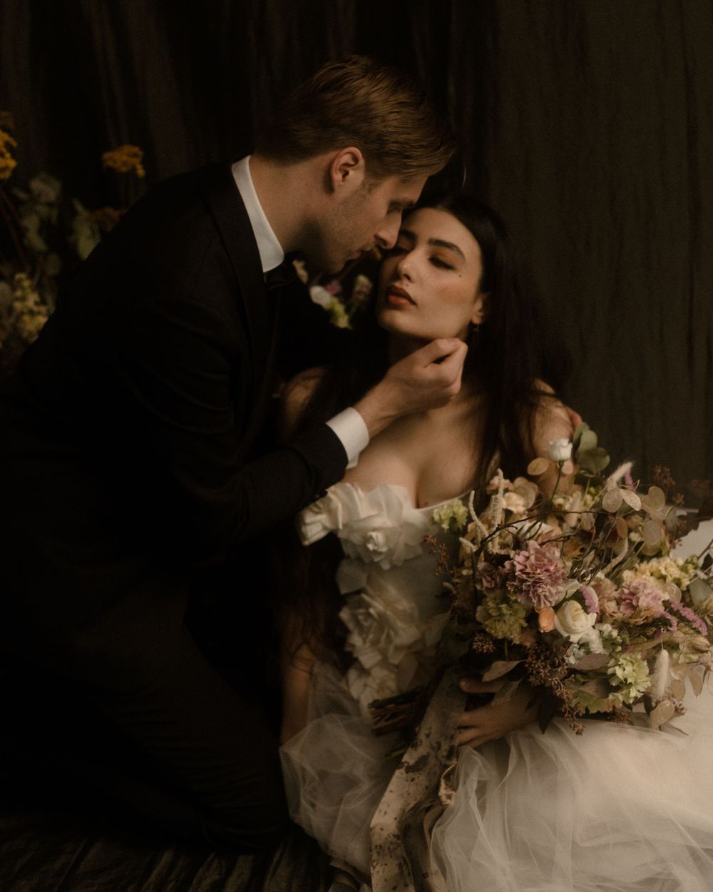 A groom in a black tuxedo tenderly holds his bride's face as she holds a lush floral bouquet in a romantic dark setting.