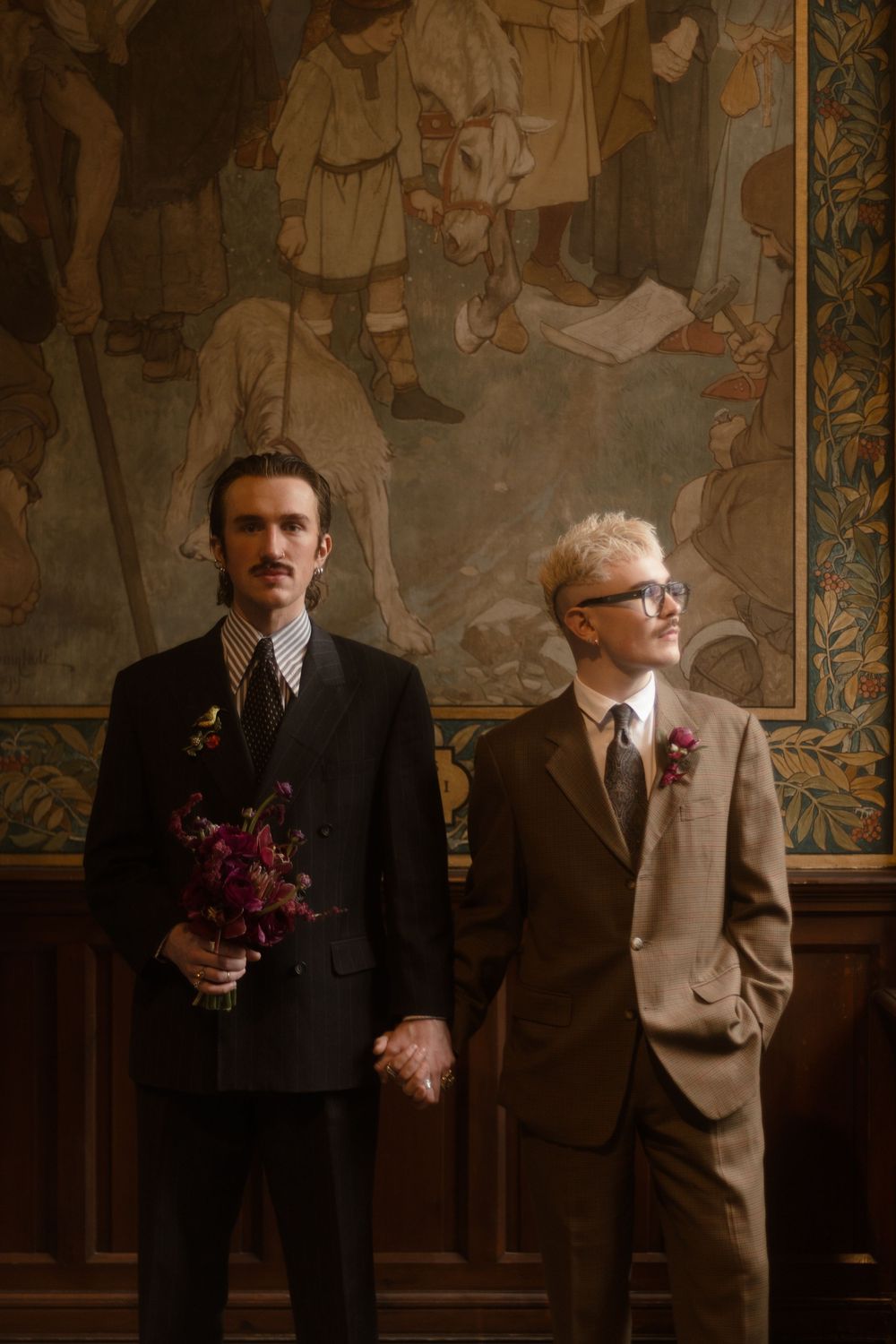 Two grooms in suits hold hands before a tapestry, one holding burgundy flowers, in an elegant wedding ceremony.