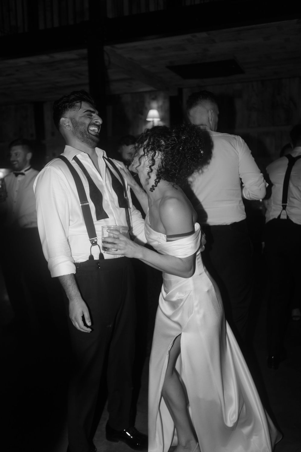 Black and white photo of a couple dancing joyfully at a wedding reception, groom in suspenders laughing with bride in elegant gown.
