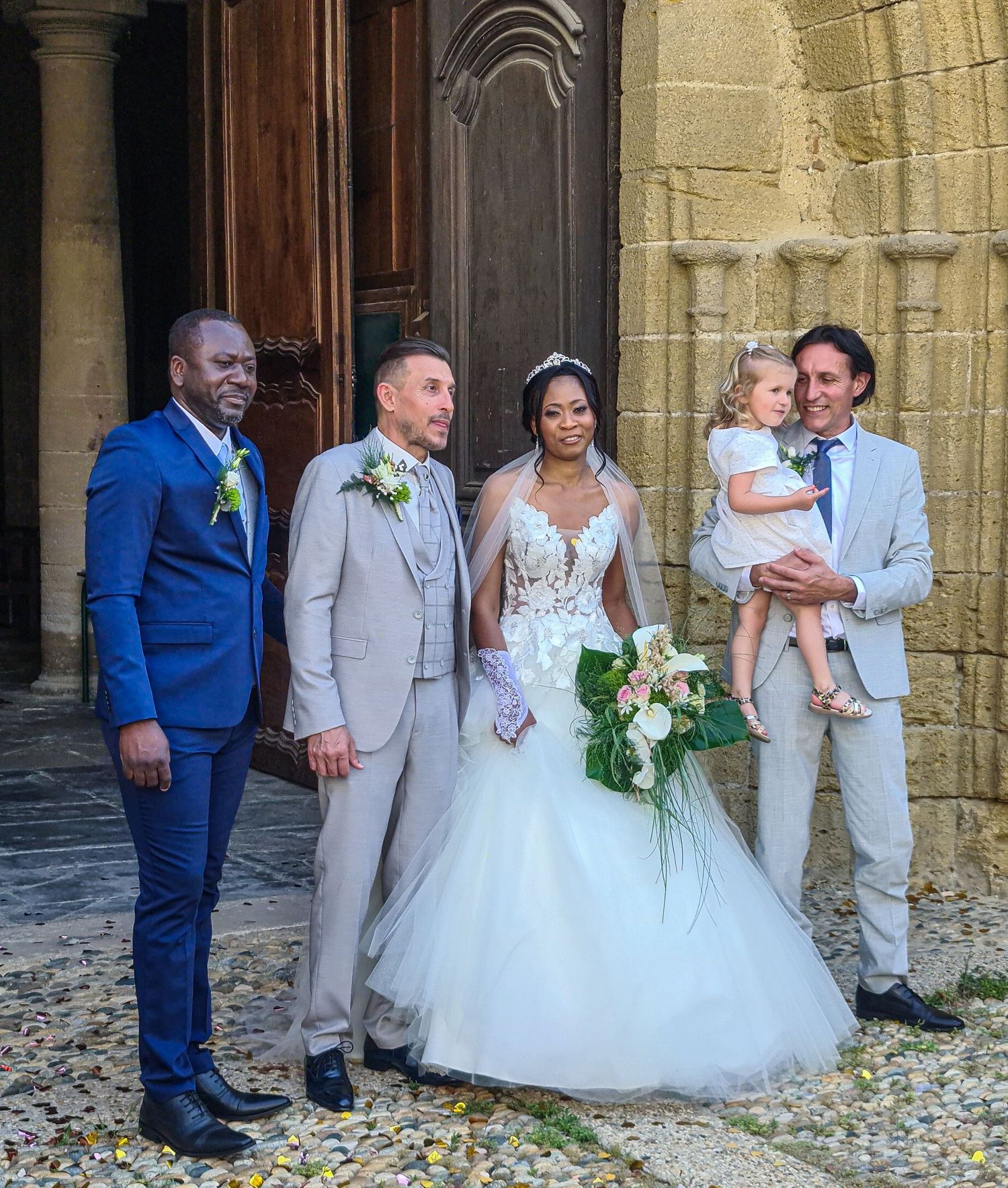 Wedding party posing outside a stone church, bride in white gown with bouquet, groom in gray suit, guests in blue and gray suits.
