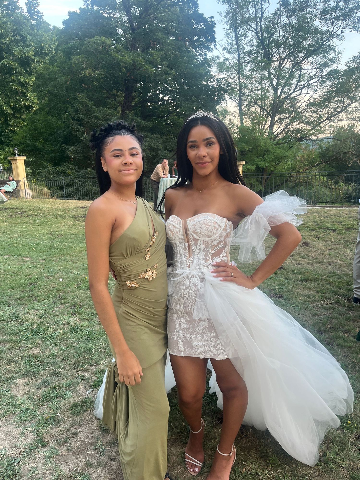Two young women posing outdoors, one in an olive green gown and one in a white lace corset dress with tulle skirt.