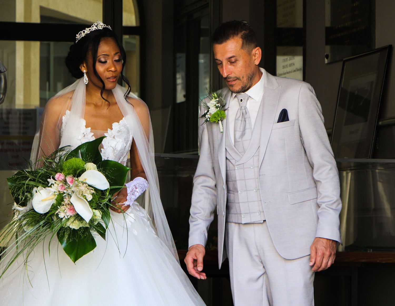 Bride in white gown with bouquet walks beside groom in gray suit at wedding ceremony.