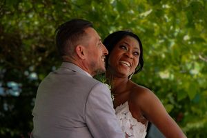 A joyful interracial couple shares a happy moment outdoors, dressed in wedding attire beneath lush green trees.