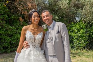 A bride in a white lace gown and groom in a gray suit pose together outdoors on their wedding day.