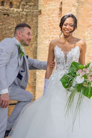 Groom in gray suit kneeling beside smiling bride in white lace gown holding floral bouquet at outdoor wedding venue.