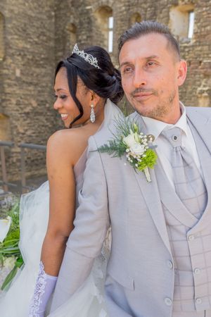 Interracial wedding couple posing outdoors, bride in white gown with tiara, groom in gray suit with boutonniere.