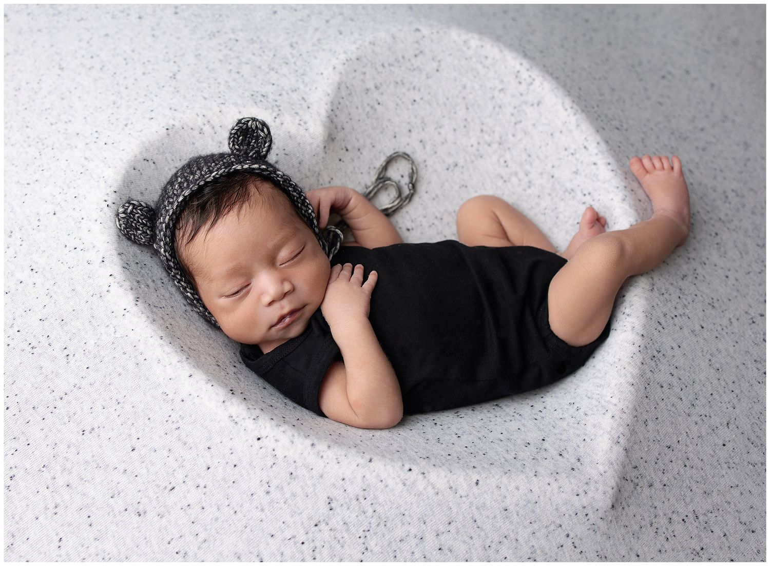 newborn posed in a fabric background heart shape, he is wearing a black onsie and black marled yarn bear bonnet.