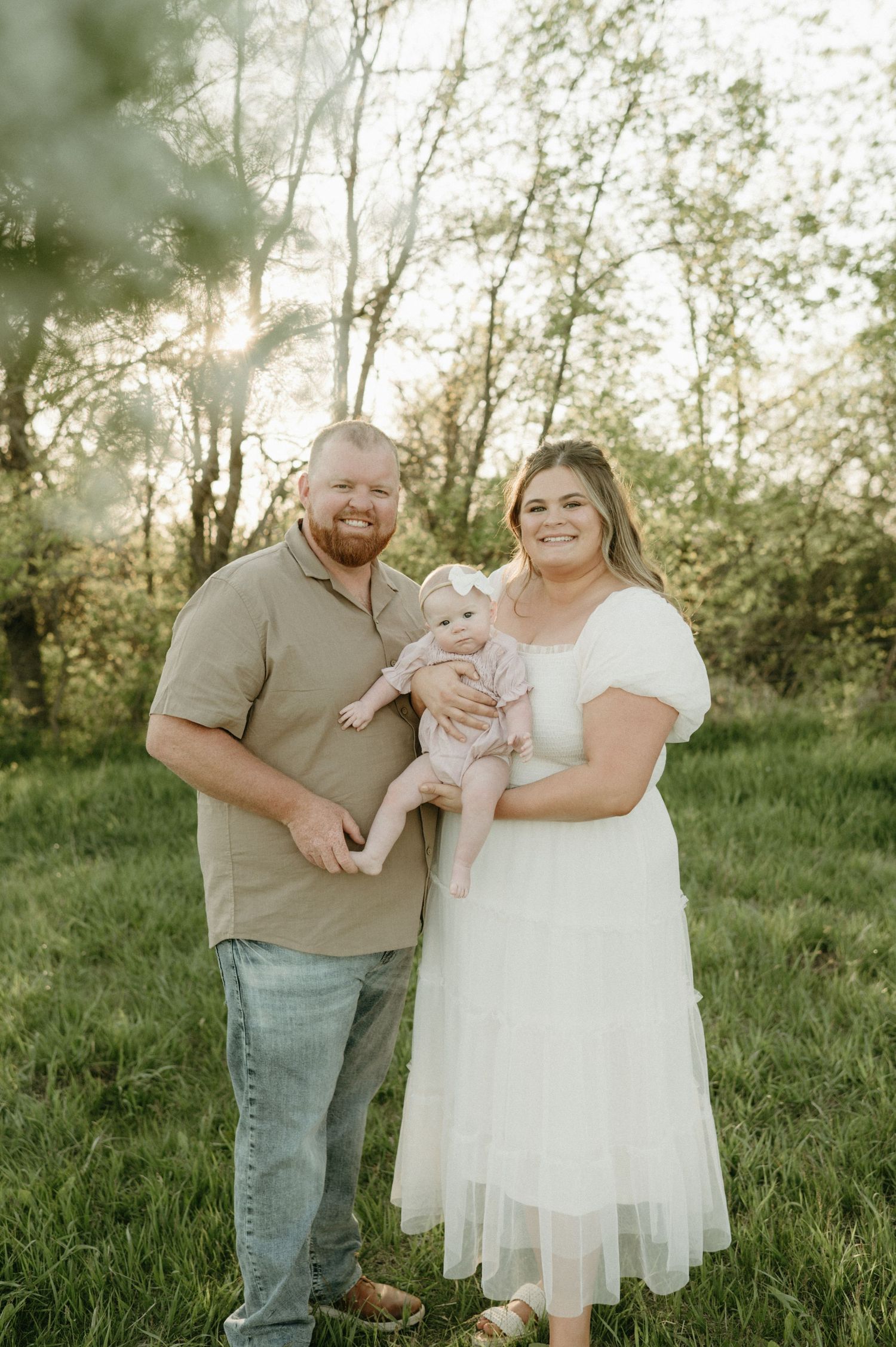 Smiling couple in green field at sunset holds their infant daughter dressed in pink between them.