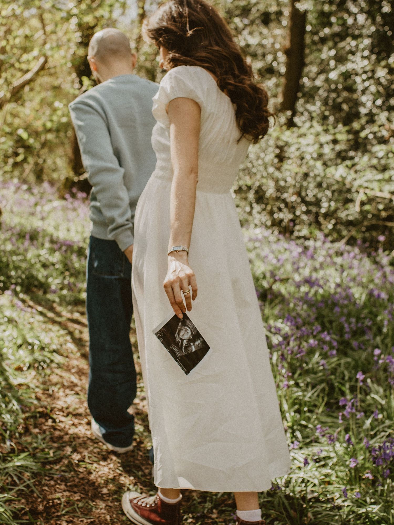 Couple walking through bluebell woods, woman in white dress holding ultrasound scan photo, pregnancy announcement.