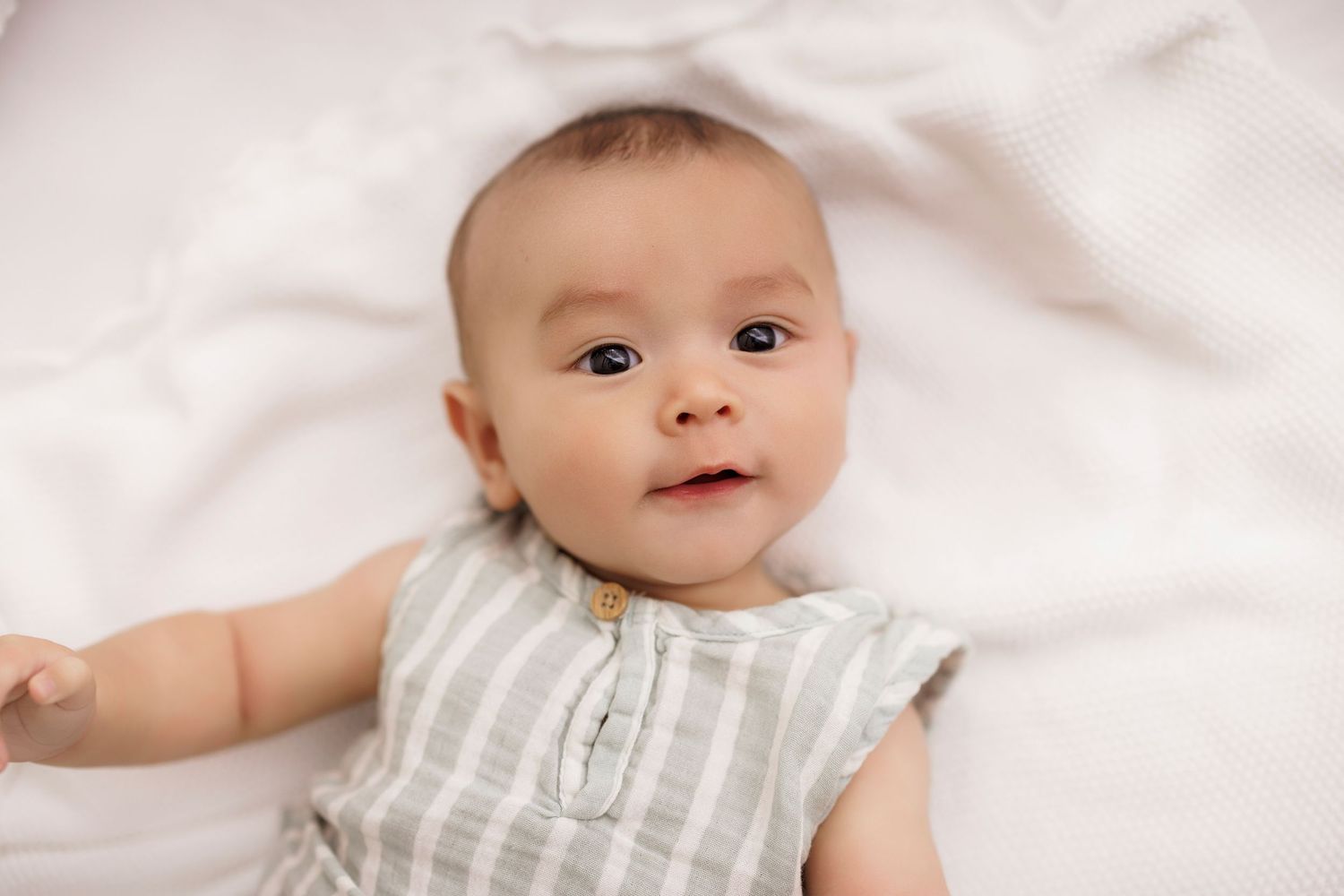 Adorable baby in striped romper lying on white blanket, looking up with curious wide eyes.
