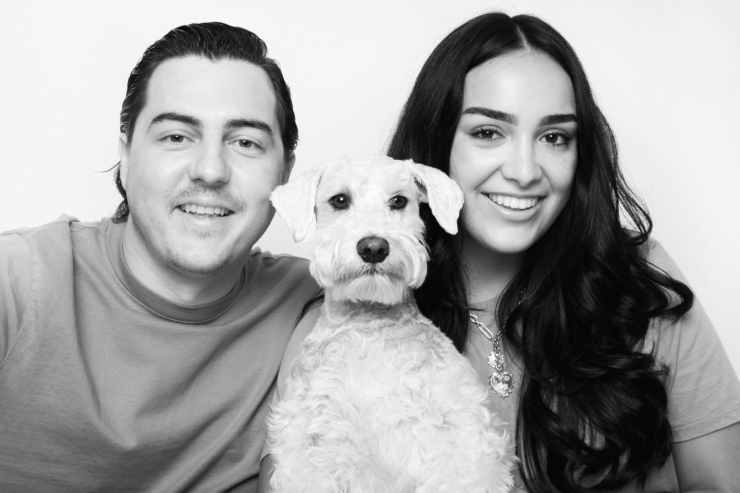 Black and white photo of a smiling couple posing with their schnauzer in a studio portrait.