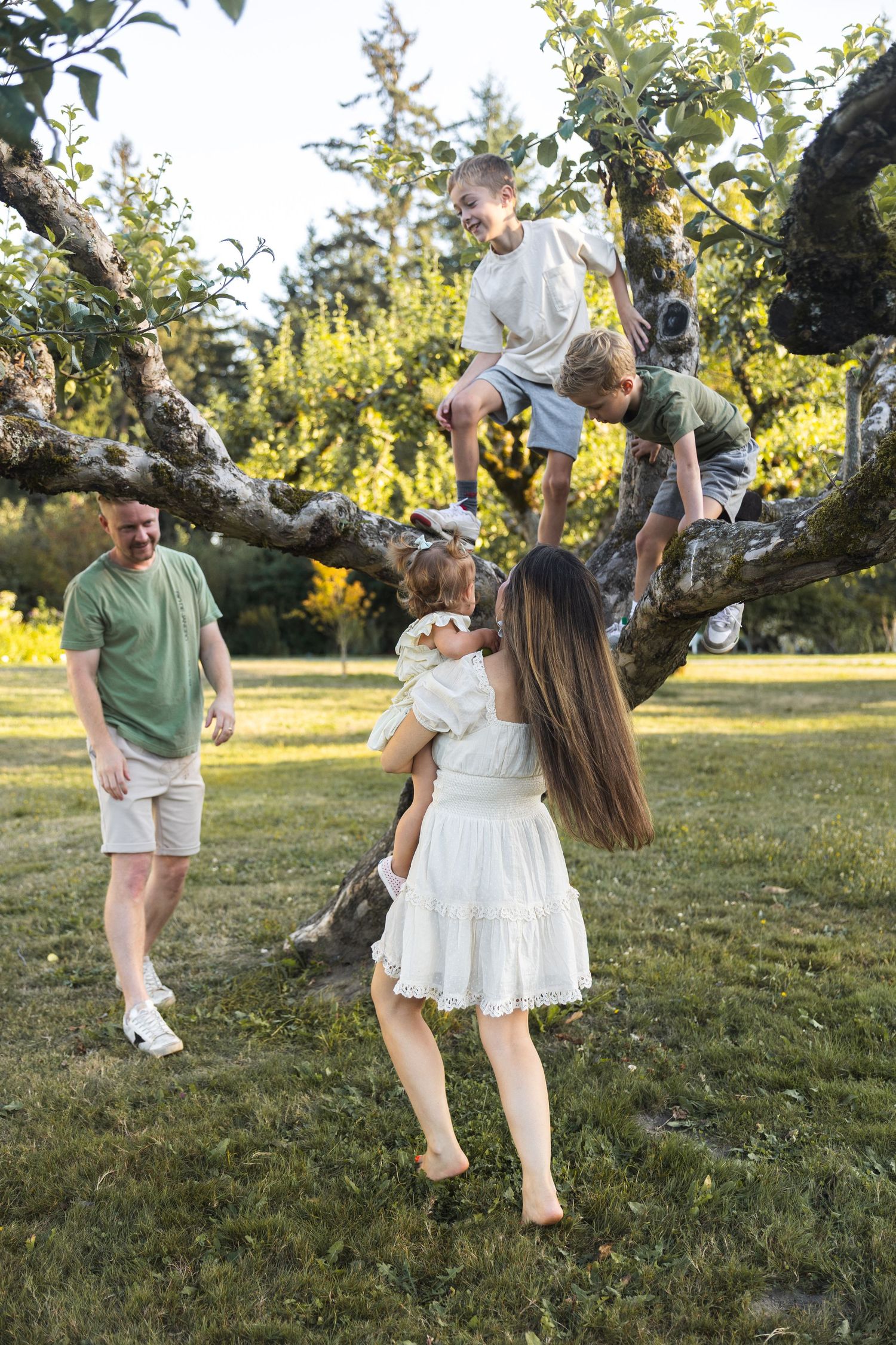 Family enjoying sunny day outdoors, two kids climbing tree while parents watch and hold toddler in green park.