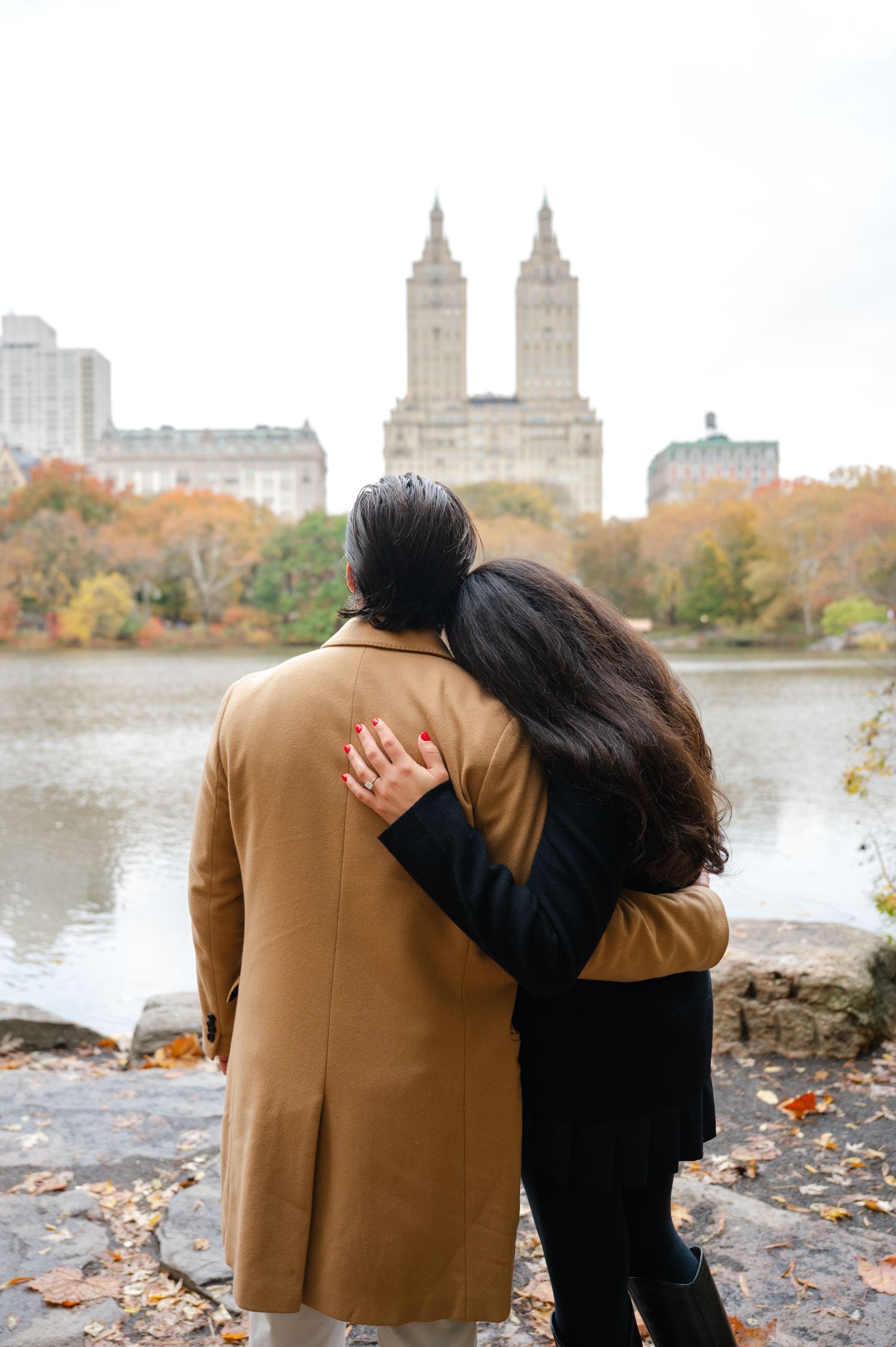 Couple embracing near the Bow Bridge in Central Park during autumn with colorful fall foliage, the lake, and San Remo in the background.