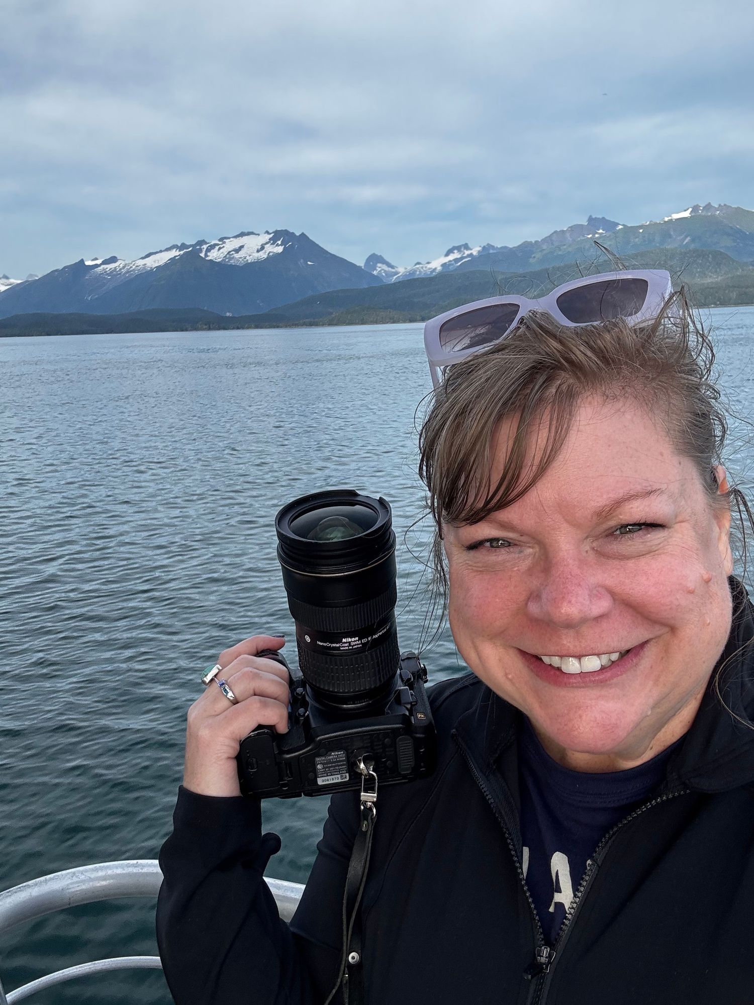 A photographer takes selfies with a camera against a scenic mountain lake backdrop in Alaska.