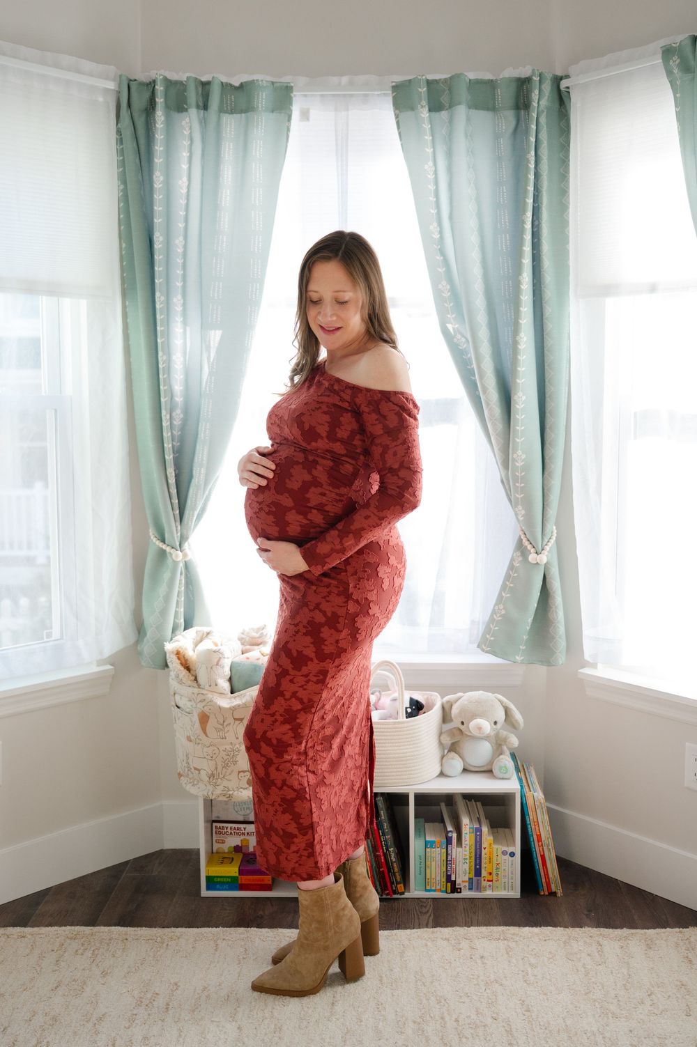Pregnant woman in red floral dress holding baby bump standing by teal curtained nursery window.
