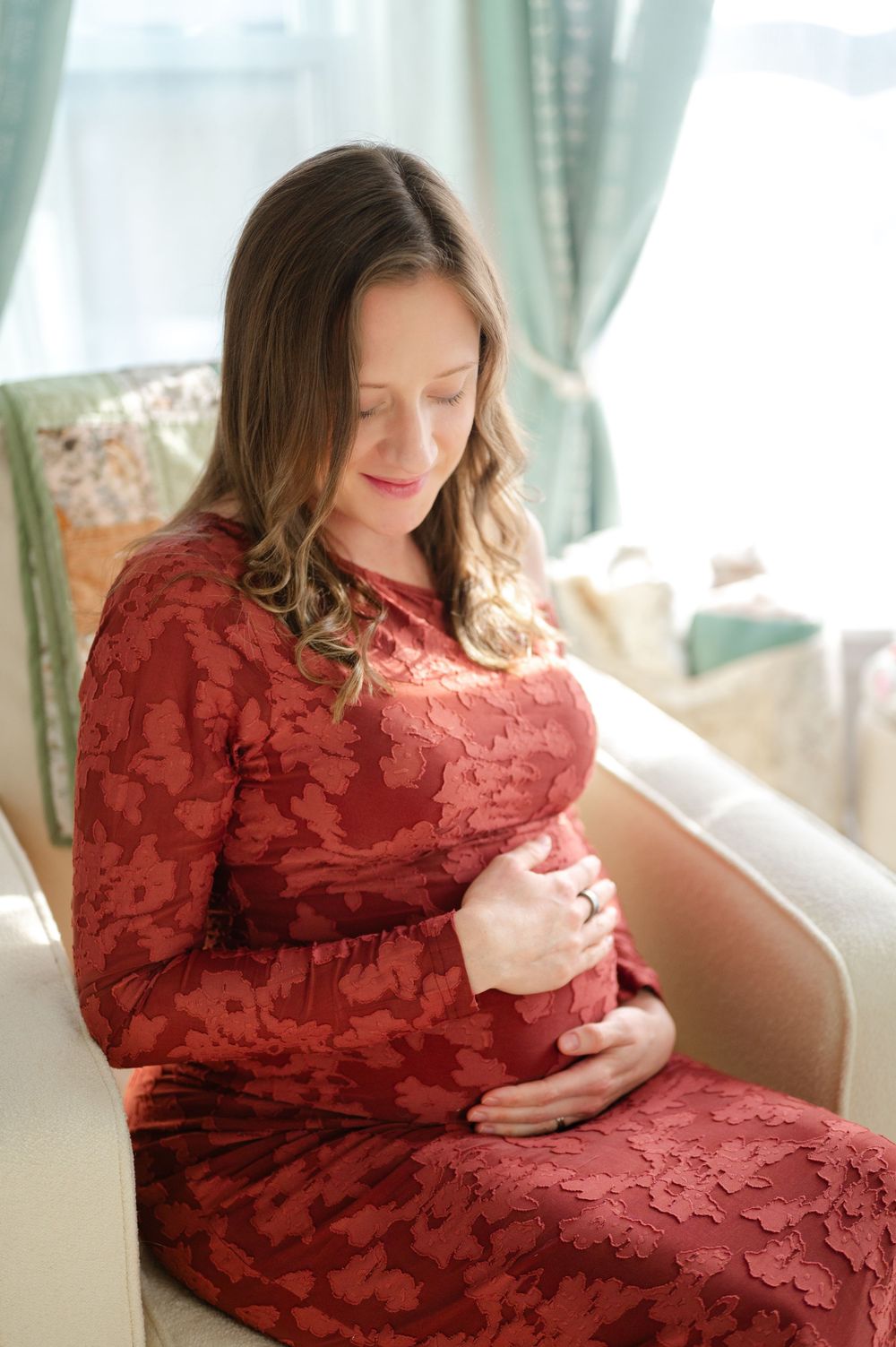 Pregnant woman in red lace dress cradling baby bump while seated in a chair by a window.