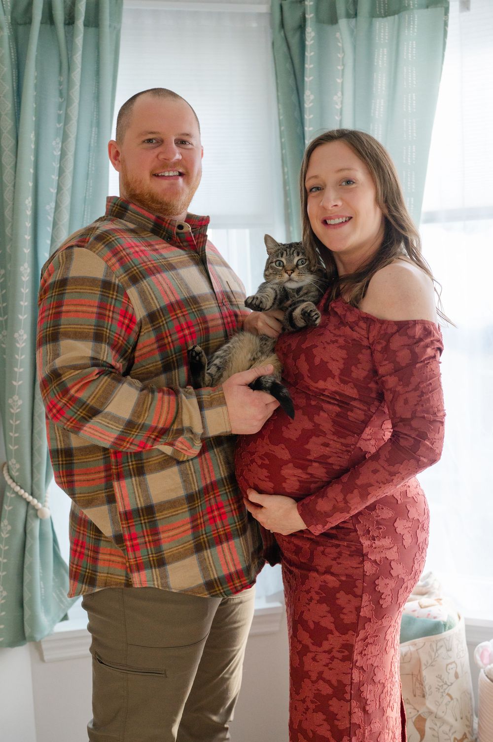 Happy expecting couple holding tabby cat, man in plaid shirt, woman in red off-shoulder maternity gown.