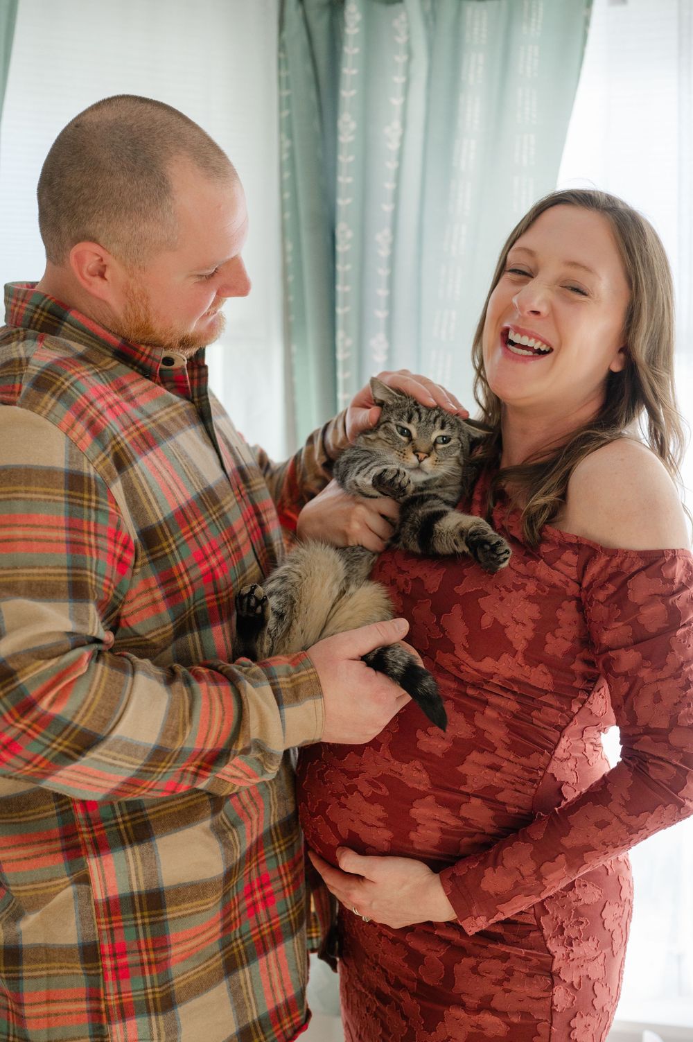 Happy expecting couple laughing while holding tabby cat in decorated home interior.